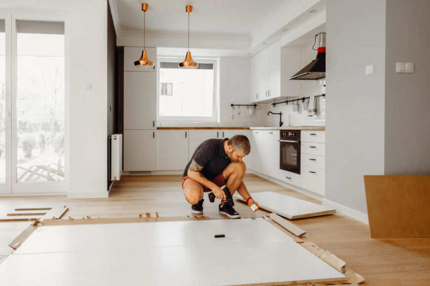 Man installing wooden flooring in a bright, modern kitchen with white cabinets, wooden countertops and copper pendant lights