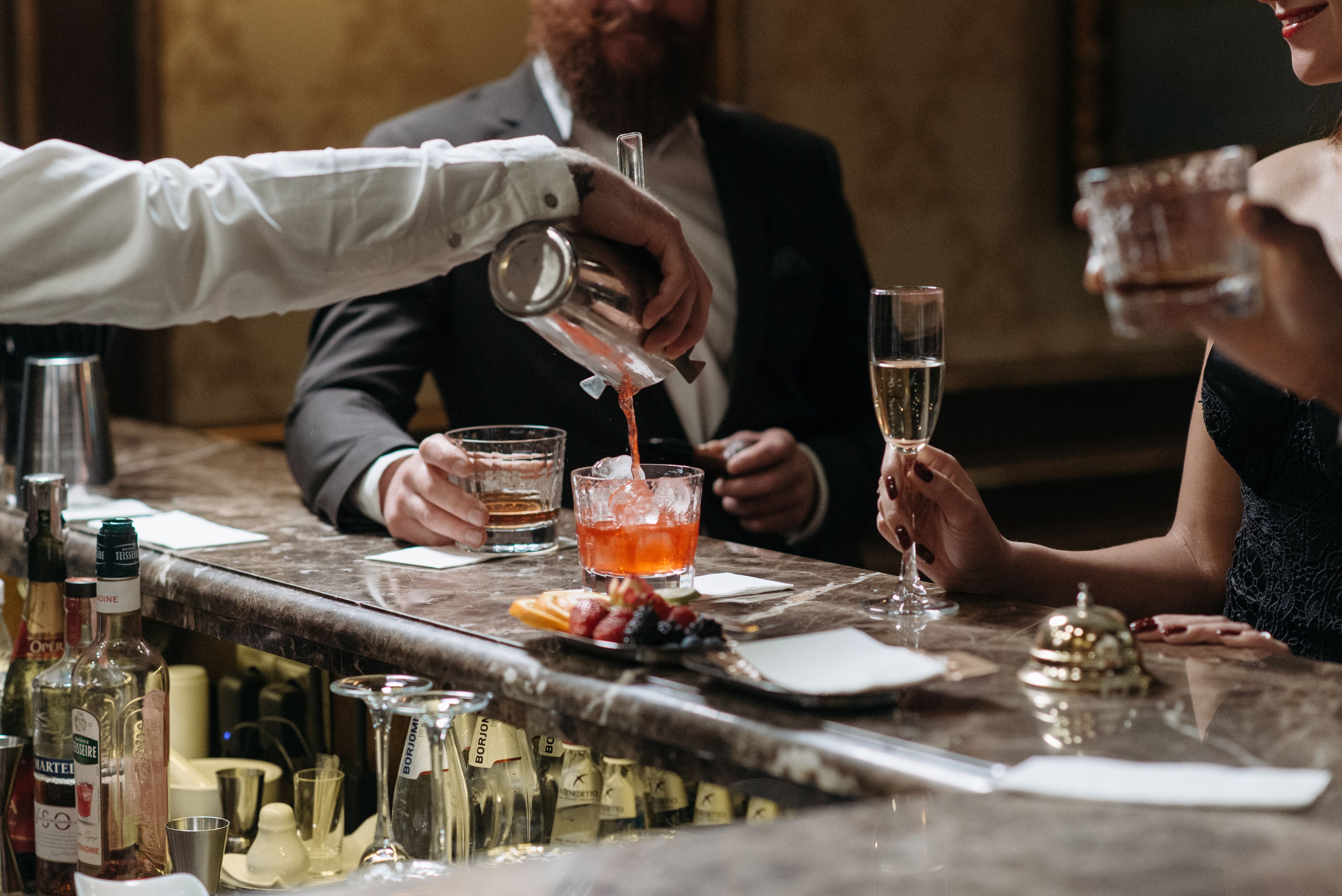 A bartender serves cocktails to well-dressed patrons in an upscale bar, evoking the atmosphere of a private club.